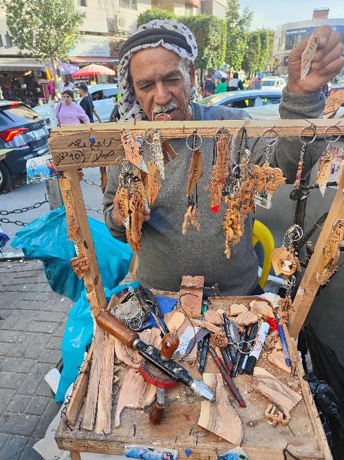 Here’s the updated description:

In the heart of Ramallah, at Al Manarah Circle, an artisan transforms pieces of olive wood into works of art. Each creation reflects Palestinian heritage, crafted with skill and dedication. Passersby can pause to watch as intricate shapes and symbols emerge from the wood, embodying both tradition and resilience. Even though I didn’t have the chance to talk to him, I wanted to share his photo and his beautiful work as a small gesture of thanks